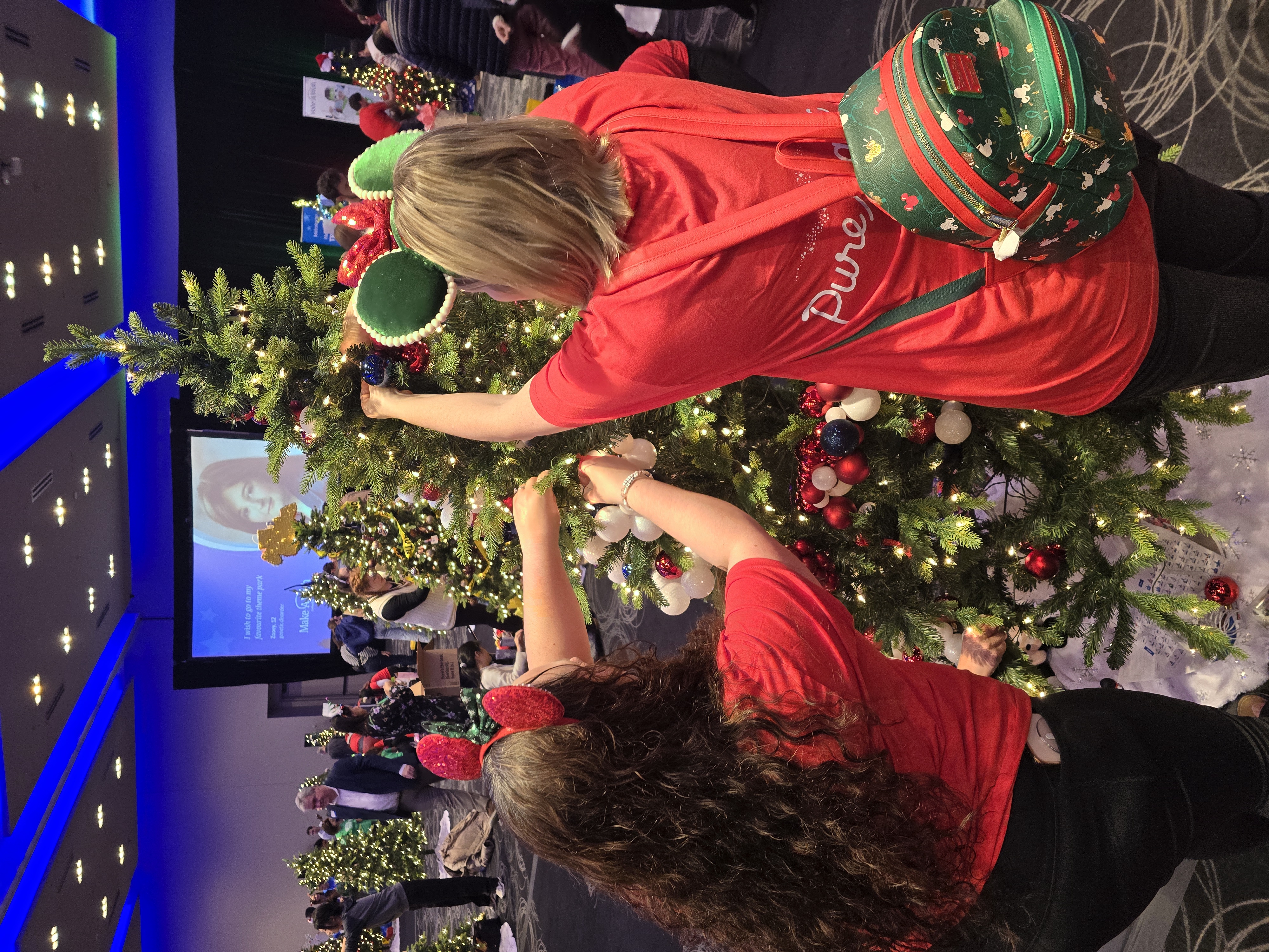 Two Pure Magic Vacations agents in red shirts hang ornaments on a Christmas tree at Trees of Joy.4_081855